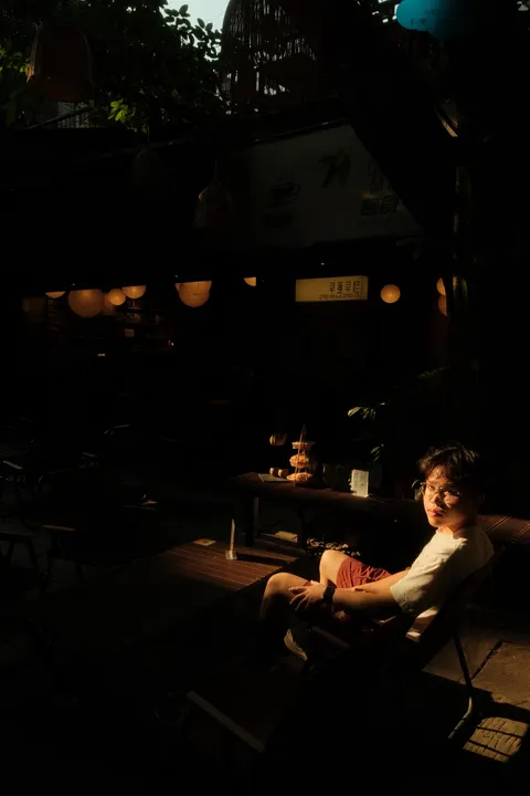 A person sits in a shadowed courtyard beneath leafy trees.