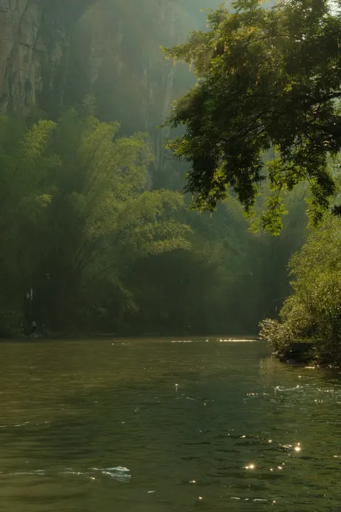 Sunlight filters through mist onto a green river and karst hills.