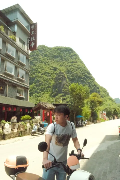 A cyclist passes village buildings at the foot of steep karst hills.