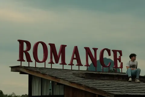 A visitor sits beneath a rooftop Romance sign overlooking green mountains.