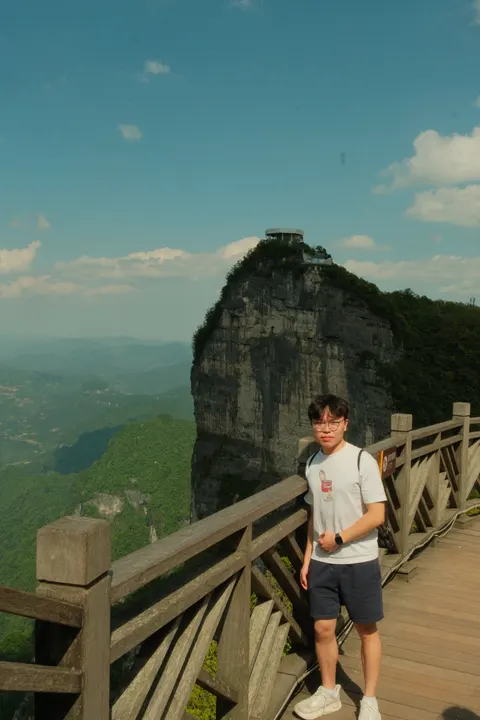 A visitor poses on a cliffside walkway above distant green mountains.