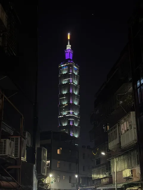 Taipei 101 glows purple above a narrow city street at night.
