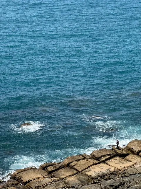 White surf breaks against dark rock in the blue Pacific Ocean.