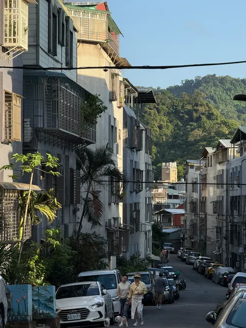 Parked cars and scooters fill a narrow street beneath green hills.
