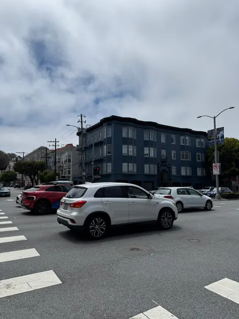 Cars cross before a dark apartment building under a cloudy sky.